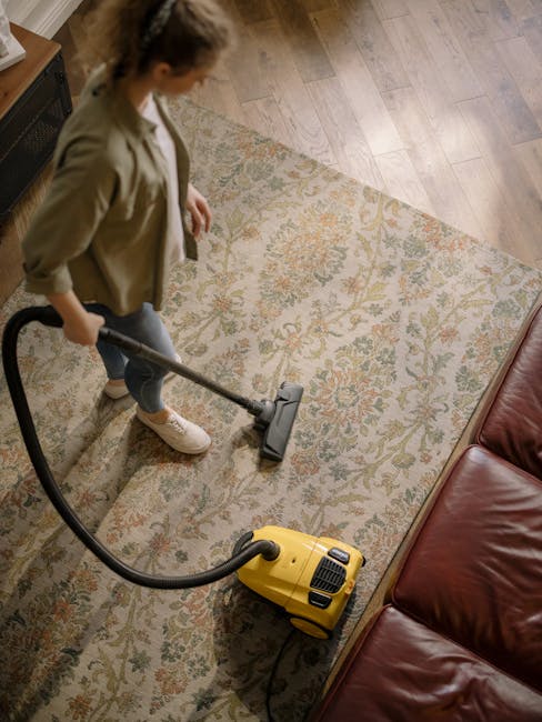 A woman is using a yellow vacuum cleaner to deep clean a patterned area rug in a living room. The rug features a floral design with shades of beige, green, and orange, placed on a wooden floor. She is
