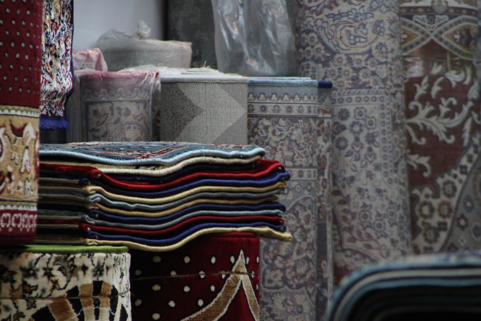 A woman is using a yellow vacuum cleaner to deep clean a patterned area rug in a living room. The rug features a floral design with shades of beige, green, and orange, placed on a wooden floor. She is